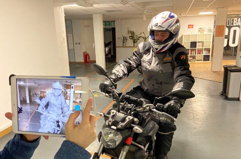Person on a motorcycle indoors while another person holds a tablet showing a live grey 3D scan of the rider and bike.