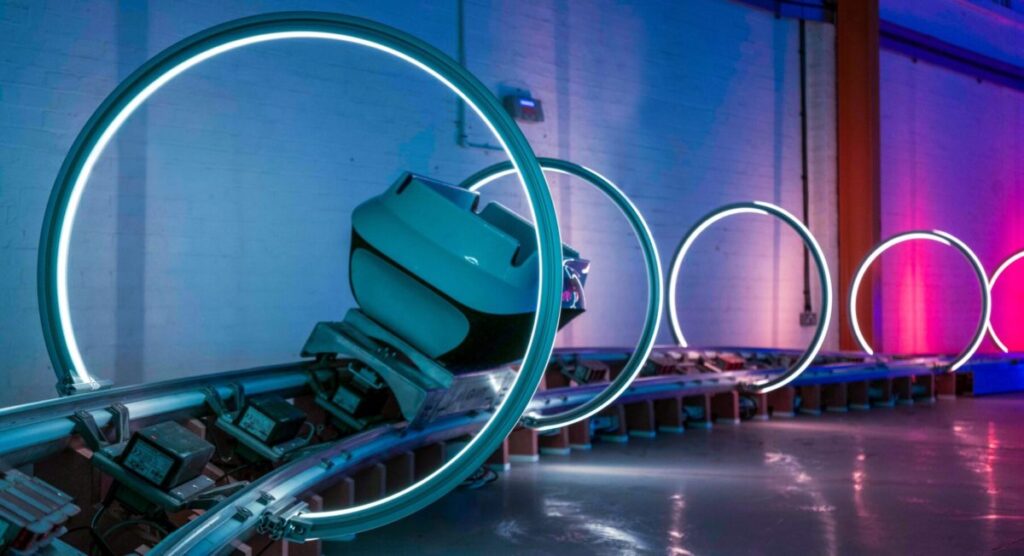 Small vehicle moving along a curved indoor test track passing through glowing circular light rings in a blue and pink lit industrial space.