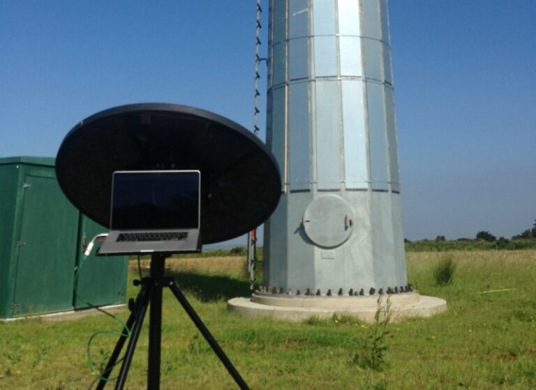 Field measurement scene showing a parabolic acoustic camera mounted on a tripod with a laptop in the center, positioned near the base of a large wind turbine tower in a grassy field under a clear blue sky.