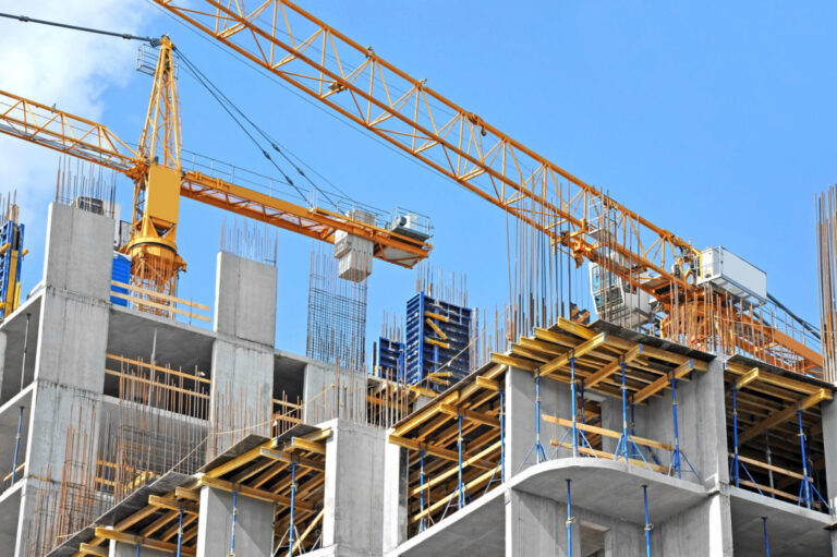 Multi-storey concrete building frame with exposed rebar and scaffolding, surrounded by large yellow tower cranes against a blue sky.