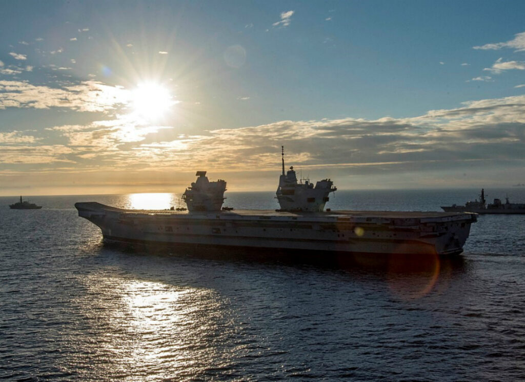 Large aircraft carrier sailing on calm open water at sunset, with the sun low over the horizon, clouds lit from behind, and smaller naval ships visible in the distance.