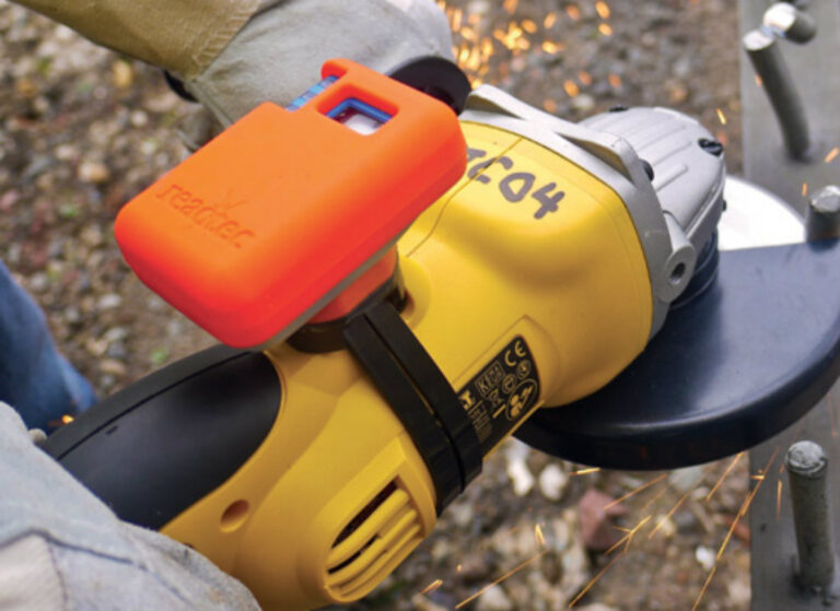 Close-up of a worker using a yellow angle grinder fitted with an orange HAVmeter device to monitor hand–arm vibration while sparks fly from the cutting disc.