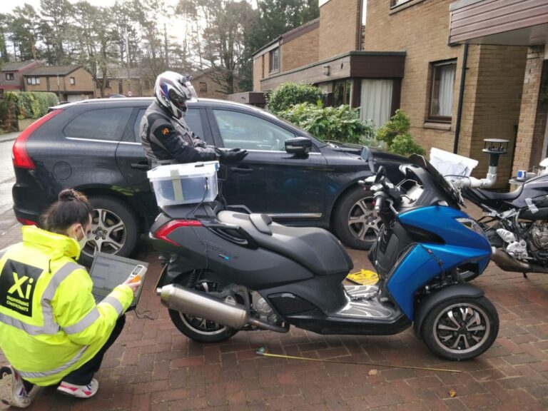 Xi engineer in a yellow hi-vis jacket recording laptop data beside a three-wheeled scooter ridden by a motorcyclist outside a residential street.