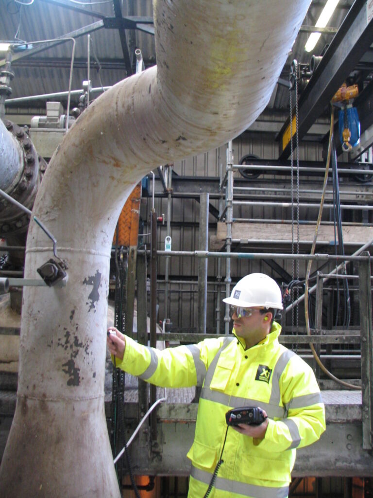 A worker in a high-visibility jacket and hard hat uses handheld inspection equipment to examine a large curved industrial pipe inside a facility.