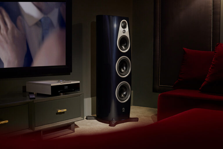 Dark home cinema room with a large floorstanding loudspeaker beside a TV and media cabinet, and a deep red sofa in the foreground.