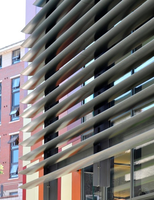 Close-up of a contemporary building facade featuring large horizontal metal louvers in front of glass windows, with a brick apartment building visible in the background.