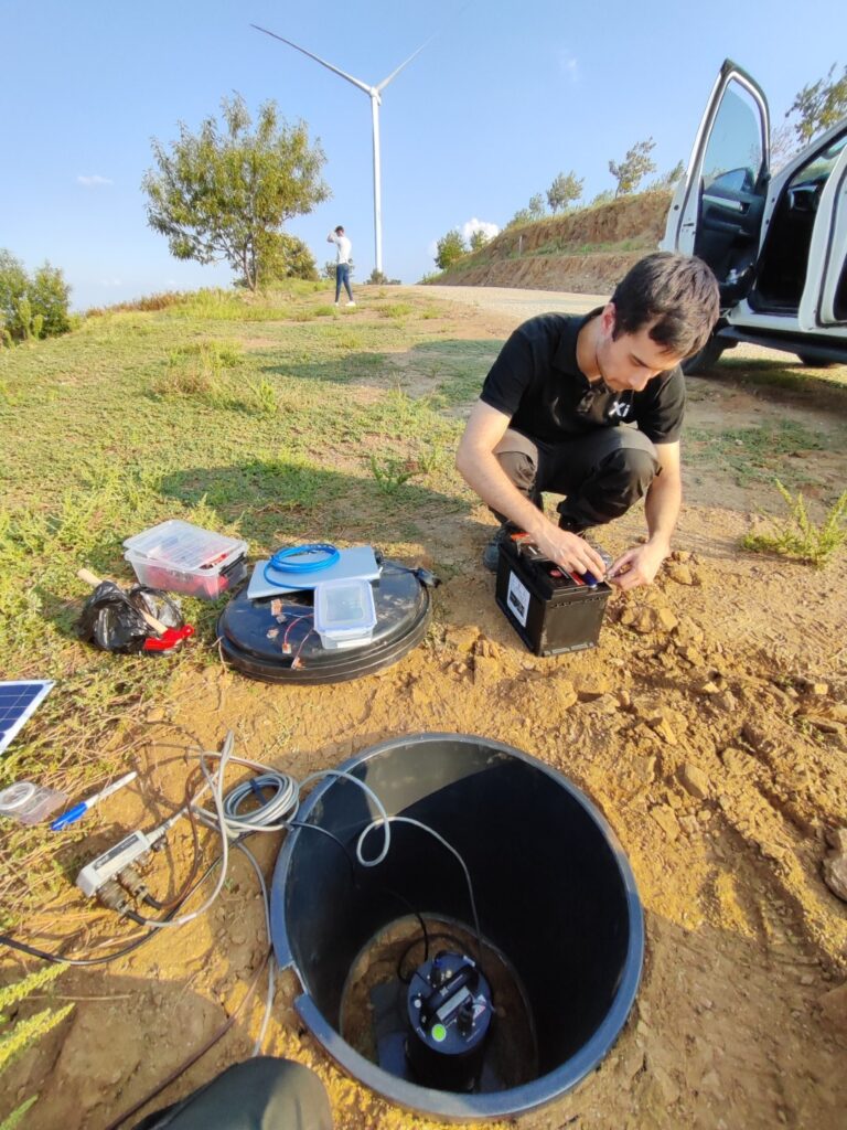 Outdoor scene showing a technician working with electronic equipment near a buried sensor enclosure, with a wind turbine and a parked car nearby.