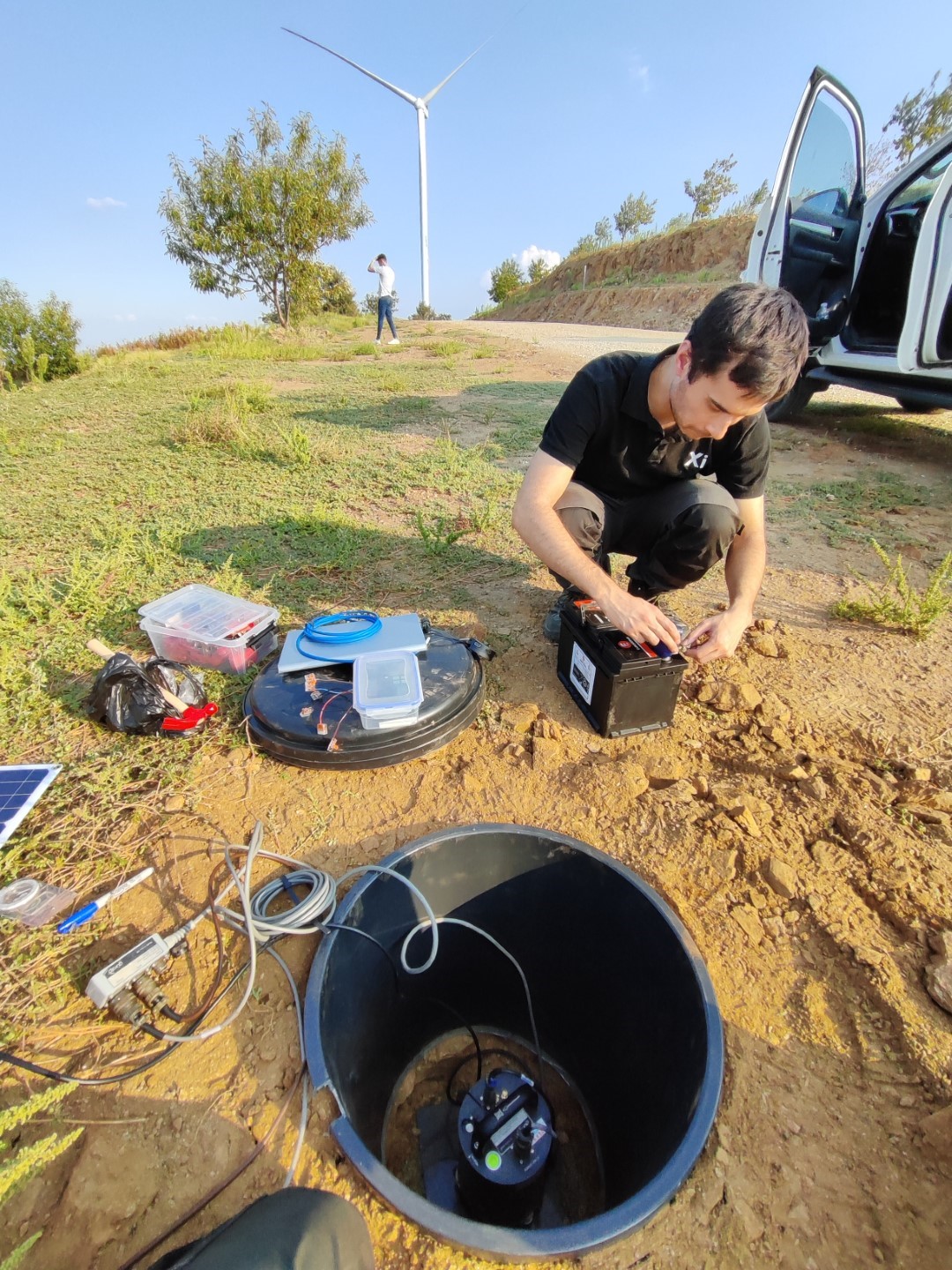 Outdoor scene showing a technician working with electronic equipment near a buried sensor enclosure, with a wind turbine and a parked car nearby.