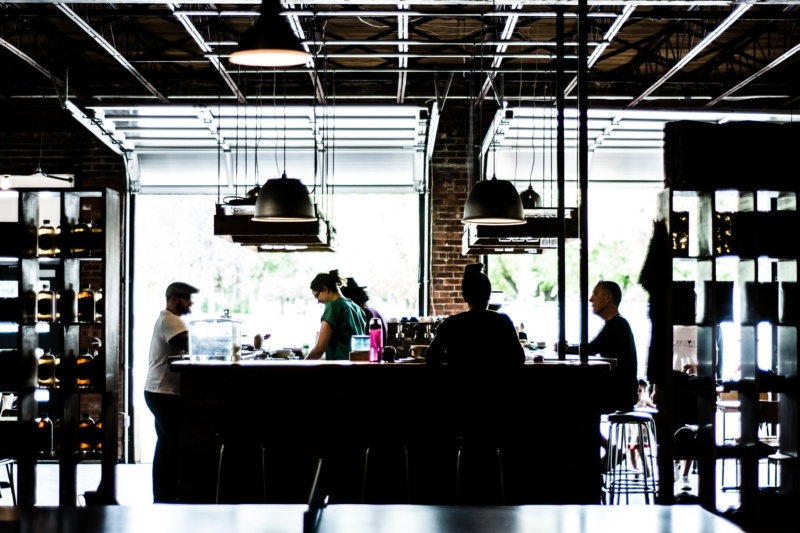 A dimly lit, industrial-style café with several people gathered around a central bar counter, including baristas preparing drinks and customers seated on stools, with large windows letting in natural light from behind.