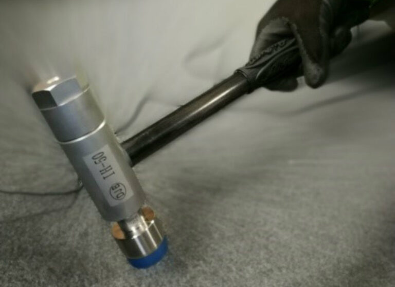 Close-up of a gloved hand holding a metal impact hammer with a blue-tipped head resting on a grey surface, used for structural dynamics measurements.