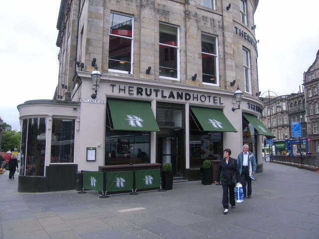 Street-level view of The Rutland Hotel on a stone corner building with green awnings, outdoor seating barriers, and pedestrians walking past on the pavement in an urban setting.