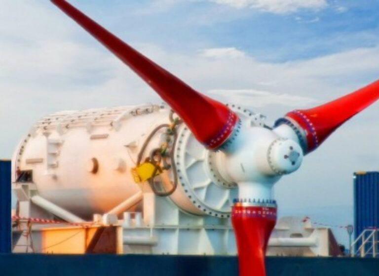 White tidal turbine nacelle with three bright red blades mounted horizontally on a quayside structure with containers in the background.