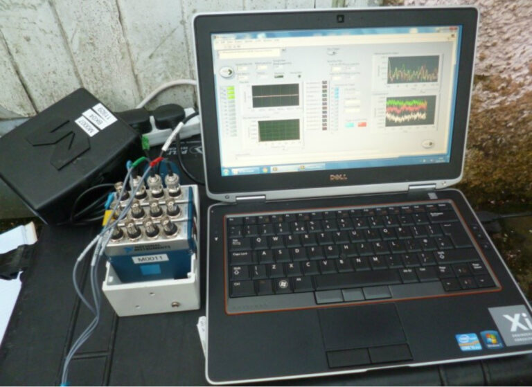 Dell laptop running vibration monitoring software connected to a compact data acquisition unit with multiple cables and BNC connectors, set up on an outdoor surface next to a wall.