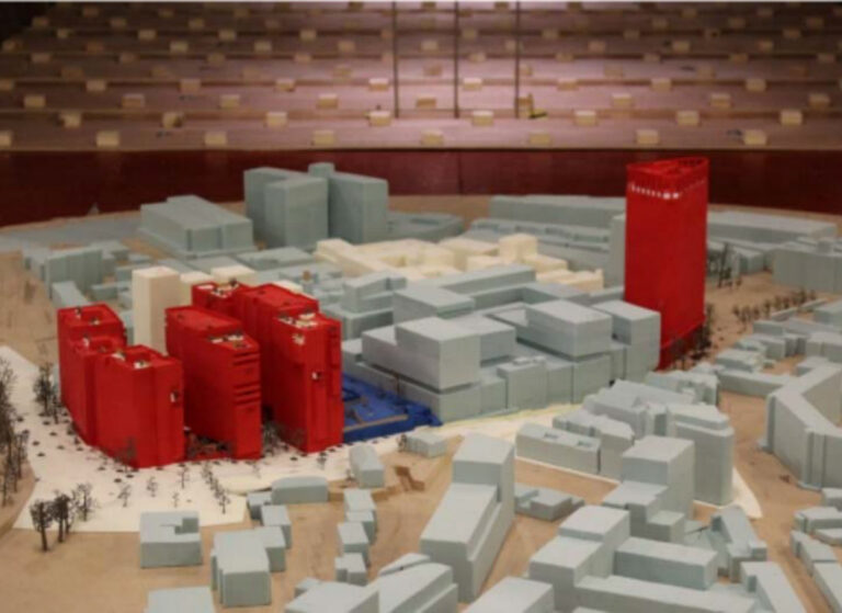 Physical scale model of an urban district showing pale blue and cream building masses, with several bright red towers and blocks indicating a proposed development area.