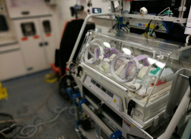 Close-up of a neonatal transport incubator secured inside an ambulance, showing the clear plastic enclosure, monitoring equipment, and medical cables ready for moving a premature or sick baby safely.