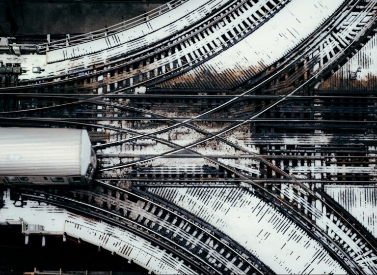 Overhead view of multiple intersecting railway tracks covered in patches of snow, with a silver train entering from the left side of the frame.