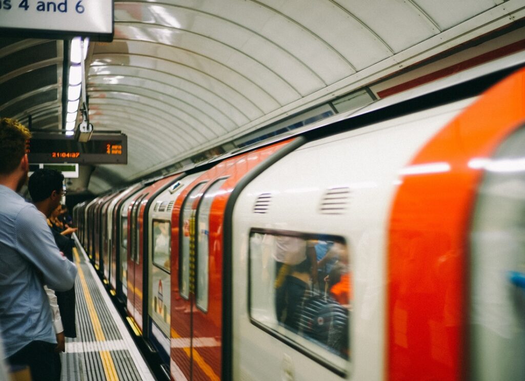 People standing on a curved underground station platform beside a red-and-white train, with doors open and passengers visible through the windows.
