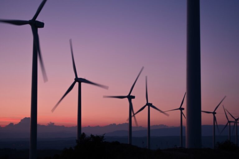 A row of wind turbines silhouetted against a colorful sunset sky, with soft purple and orange hues and distant hills in the background.