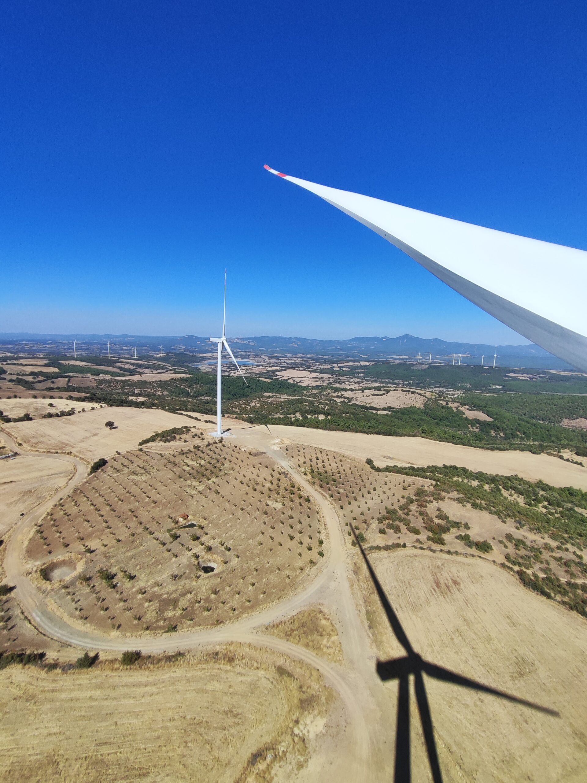 View from a wind turbine blade over a dry rural onshore wind farm with turbines, access roads and hills in the distance.