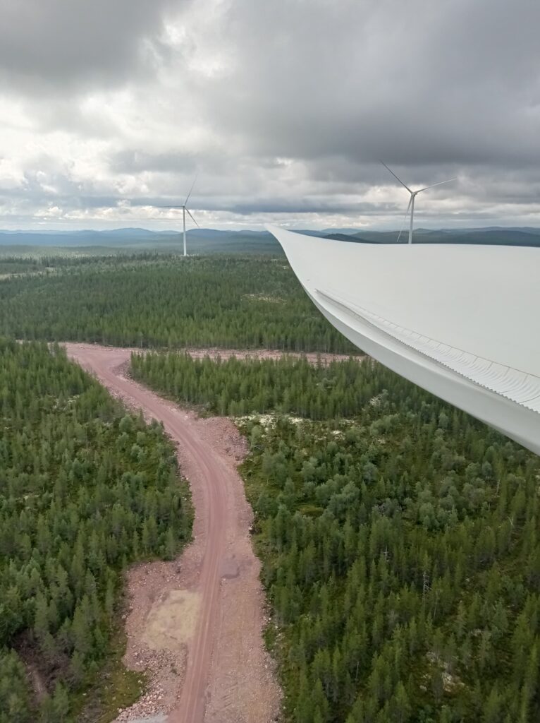 View from a wind turbine blade over a forested onshore wind farm with access road and turbines in the distance.